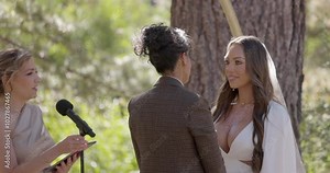 In a charming outdoor setting, a beautiful wedding ceremony highlights two diverse young women in stunning gowns, joyfully celebrating their deep love and commitment to one another