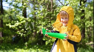 Little boy in yellow raincoat plays with sprayer in summer park