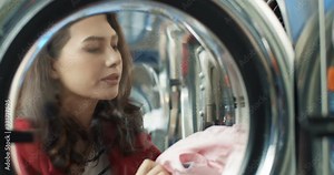 Close up of pretty Caucasian woman opening washing machine and taking out clean clothes after wash. Beautiful stylish young girl laying out clothing in laundry service room.