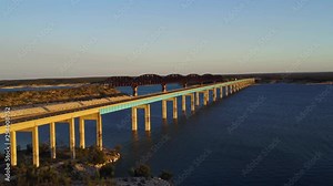 Aerial of Bridge over Lake Amistad outside Del Rio Texas on the United States - Mexico border during Golden Hour. Fishing Boats and Semi Trucks pass by and train tracks run parallel to highway.