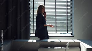 Angry woman in stylish suit shouting and gesturing emotionally during mobile conversation with business colleague while standing near window. Sunlight coming at dark office through opened blinds.