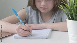 Close up kid's girl hand making notes in notebook at desk. Student child girl holding pen doing homework at home, or doing examination at school, education, distance studying concept