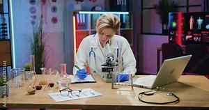Attractive confident observant skilled blond woman scientist biologist in lab coat and gloves watching on samples under microscope and noting details into paper report