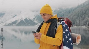 American traveler man using smartphone in hiking winter tour. Snowy lake and mountains against background. Traveling and communication concept: hiker scrolling and tapping on line by cell phone