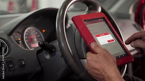 Auto electrician uses OBD-II scanner to diagnose car issues. Expert checks vehicle diagnostics, fault codes on tablet in workshop. Maintenance, service, repair process shown hands-on.