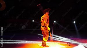 A clown with a funny hairstyle entertains guests in the circus arena in the light of colorful spotlights and smoke. He's tap dancing. Circus performance.