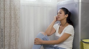 housewife sitting back against the washer is feeling bored, and letting out sigh, touching face while waiting for laundry.