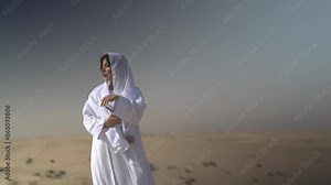 A woman in a long dress walks through the sand dunes of a desert. The expansive landscape under a clear sky highlights the serene and solitary nature of the scene.