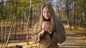 Front view of stylish, young lady walking in park, drinking coffee. Attractive woman standing, smelling coffee, enjoying, looking at camera. Concept of modern lifestyle.