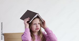Little girl puts thick book on head playfully blowing air. Bored child demonstrates unwillingness to perform homework leaning on table