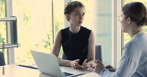 Serious pretty young employee woman talking to business colleague at workplace, discussing project, online app for job at laptop. Female mentor teaching intern, telling about work tasks Generative AI