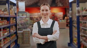 Shop assistant stands in supermarket smiling. Young red-haired woman shows and advertises groceries on racks looking straight at buyers closeup