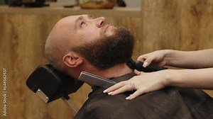 The barber takes care of the client's beard using cosmetic devices for cutting and professional care. Brutal dark interior. Professional young female hairdresser-stylist working with a client. Stock Video