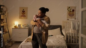 Medium full shot of Hispanic mother in casual pyjamas and cardie holding fussy baby son, walking around room, swaying and rocking to calm him down for daytime nap