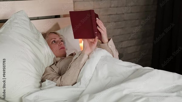 Late evening, a woman reads a book while lying in bed in her bedroom.