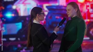 Female presenter reporting on air outside. Woman news host journalist taking interview with young female on the street, police and ambulance cars at the back.