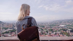 Young blonde woman traveler with backpack observing urban development of Tbilisi, Georgia from hill height, showing unique architecture of city and winding Kura River. Rear view.