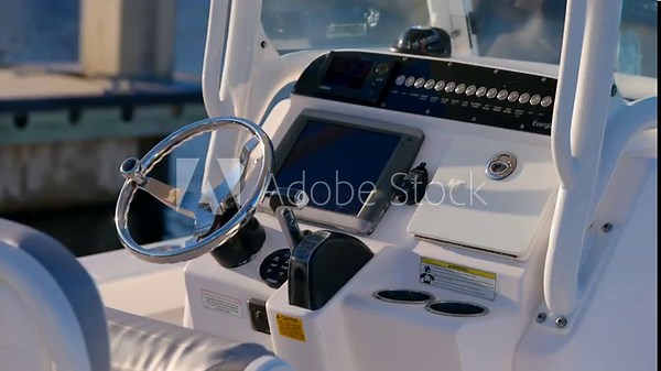 Console of motorboat shows wheel, screen, and buttons. Dock and water are seen behind. Labels give warnings, switches control parts, everything looks ready to use.