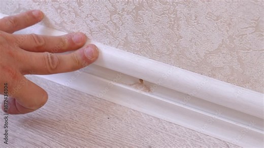 Close-up view of a professional worker drilling a hole in the wall, inserting a dowel, and screwing a white plastic baseboard to the wall, completing the installation process