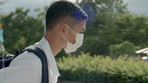 An Asian male student in white high school uniform wearing the mask stand on the side of the road waiting for car to go back home after school during the Coronavirus 2019 (Covid-19) epidemic.