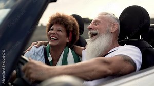 Trendy senior couple having fun inside convertible car - Multiracial mature people on a road trip with cabriolet car