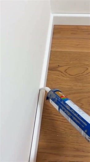 A person applies white sealant along baseboard edge beside wooden floor #construction #popular