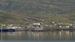 Shot of a small yacht sailing out of the harbour at Castlebay on a sunny day. The ancient Kisimul Castle is visible as well as a church. Filmed on the Isle of Barra, part of the Outer Hebrides.
