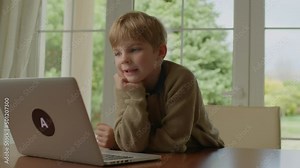 School boy talking using laptop sitting at the table by the window. Blonde kid learning online alone at home.