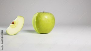 Fresh tasty green apple piece falling down and rolling onto its side near the apple on dry white floor. Shooting with high-speed camera in slow motion mode. White background isolated.