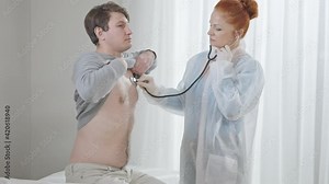Concentrated woman listening internal sounds as man sitting on medical couch in hospital ward. Caucasian doctor using stethoscope for chest auscultation as patient inhaling and exhaling.