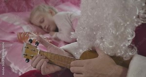 Close-up of unrecognizable Caucasian man in Santa Claus costume with false beard playing ukulele as little blond girl sleeping at the background. Cinema 4k ProRes HQ.