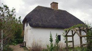 Thatched-roof cottage with chimney, surrounded by lush greenery and a wooden trellis, evokes rural charm. A serene and traditional english village country home scene