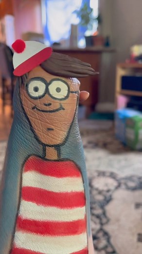 Where oh where is Waldo?!?!? 🥰😂 #cosplay #whereswaldo #limbdifferenceawareness #makeyousmile Video description: Alexis is sitting at her living room table wearing an orange T-shirt with dinosaur skeletons on it. There is a wooden bookshelf covered in plants behind her. Her brownish reddish greyish hair falls in layers around her shoulders. She is using red and white paper, glue, red felt, brown clay, red, black, white, brown and beige paint to transform her stump into Waldo from Where’s Waldo.