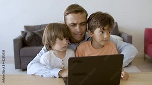 Serious dad working on laptop and embracing sons. Concentrated Caucasian father using computer and talking with kids on his knees. Front view. Fatherhood, childhood and digital technology concept