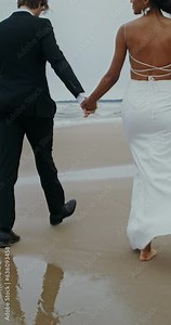 The bride and groom hold hands walking on the beach, the bride holds shoes in her hands
