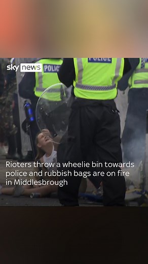 Rioters throw a wheelie bin towards police and rubbish bags are on fire in Middlesbrough. A woman can be seen being helped by police, sitting just inches from litter in flames. | Sky News