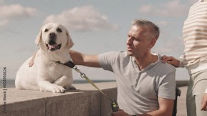Smiling man in his 50s sitting in wheelchair outdoors in summer and petting his light yellow Labrador retriever then his wife joining them and putting hands on his shoulder