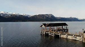 Low fly-over shot of a boathouse on the end of a pier in a calm lake. Majestic snow covered mountains paint the most beautiful background on a overcast day. Stock Video