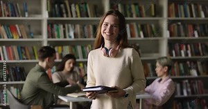 Pretty redhead girl standing in library, diverse schoolmates behind. High school student pose on bookshelves background, excellent studies in university or international college, get higher education