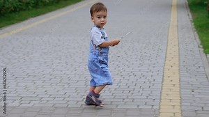 Adorable little kid in a jeans romper walking by the road. Kid holding a magazine in his hands comes up to a big statue of a cat carved from wood.
