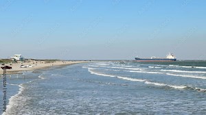 Afternoon beach scene at the Gulf of Mexico with sand, waves, cars, people enjoying themselves, and a crude oil tanker ship heading toward a refinery on a sunny day.