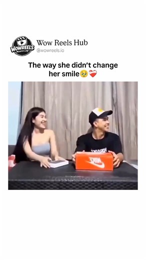 Wow Reels Hub™ on Instagram: "The video depicts a young couple seated at a table in a casual indoor setting with beige curtains in the background, engaging in a heartwarming gift-opening moment set to a romantic ballad. The man, wearing a black t-shirt and a backward cap, presents his partner—a woman with long dark hair in a strapless top—with a large orange Nike shoebox. She smiles brightly as she begins to unpack it, revealing a series of nested smaller boxes inside, each one opened with antic