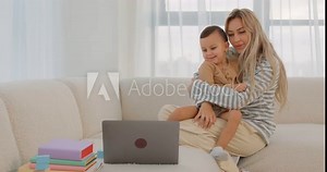 Awesome Blonde Mom With Infant Son On Knees Using PC Computer In Living Room, Mommy touching kid's nose, tickling him, has fun with adorable son Cute boy gets pleasure from spending time with parent