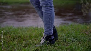 women's feet in jeans and dark boots with thick heels and white laces walk on the green grass along the river. side view