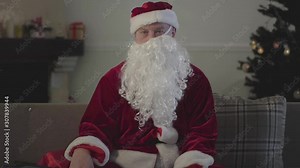 Drunk old man with false beard crooked on the face in the costume of Santa Claus sitting on the sofa at home, holding empty bottle. Bad Santa. Alcoholism, depression, negativism, loneliness