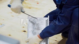 Worker man wear blue overalls, white protective gloves, squatting, hold in hand tool, use file and burnish edges of steel metal billet of cable channel on background of wooden floor. Close up view.
