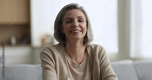 Pretty middle aged grey haired woman posing at home, looking at camera, getting from serious to positive, cheerful, happy, sitting on couch in living room, expressing emotions. Female front portrait