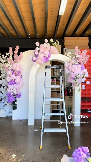 Floral arches can be used at any type of event such as a wedding, birthday or christening. Start by selecting a sturdy frame, ensuring it’s securely anchored. Choose flowers and greenery that match your theme. Begin by covering the frame with a base layer. Next, arrange larger flowers, distributing them evenly across the arch. Fill in gaps with smaller flowers and any additional flowers making sure to maintain balance and symmetry. For added dimension, let some elements drape naturally. #howto #