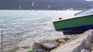 White sailboats. Green boat hull on the shore. Azure color of water. Mountains in the background. Overcast. Strong wind. Sailing. Leisure. Vacation. Water tourism.