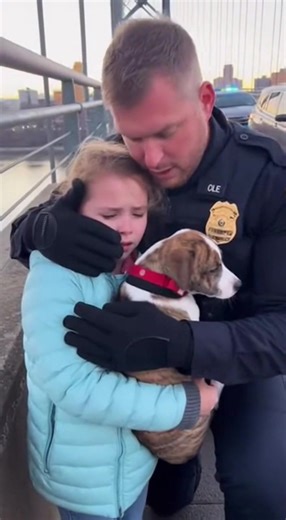“He dropped to his knees in the middle of the bridge — and suddenly no one could breathe.” Traffic stopped. Not because of an accident. Not because of sirens. But because a white male police officer in his early 40s had just knelt down in front of a little girl holding a dog at the edge of the bridge. The city froze. Wind rushed through steel railings. Cars idled. Phones hovered in midair, then slowly lowered. Even the river below seemed quieter. The officer was breathing hard, sweat darkening h